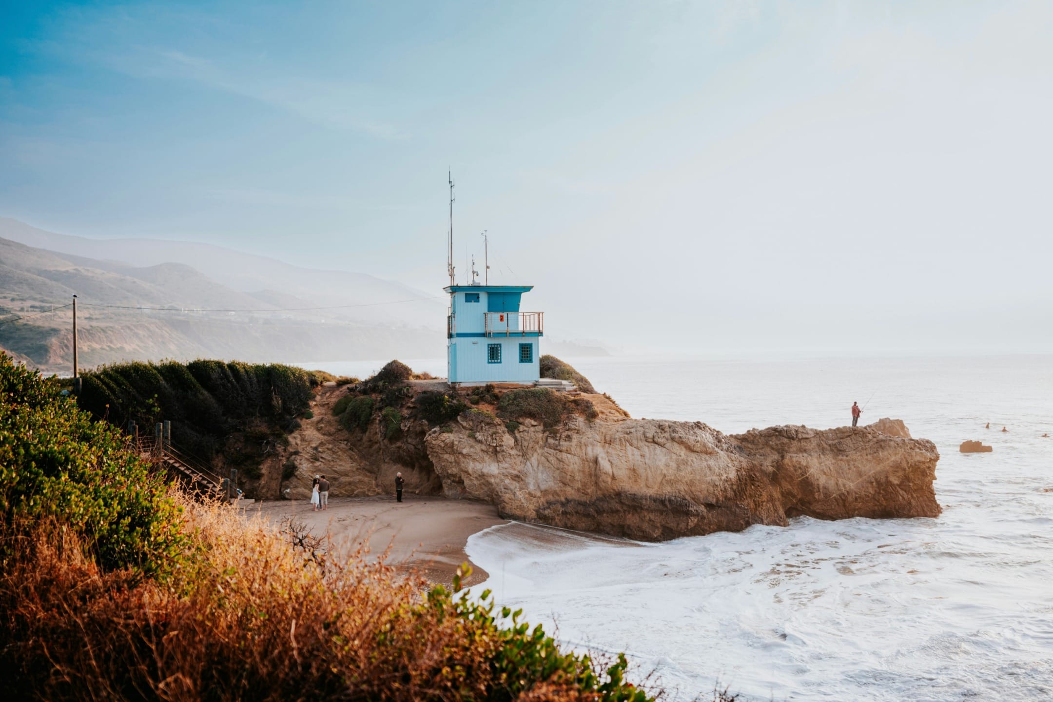 Blue lifeguard tower on a rocky cliff overlooking the ocean with waves crashing below and mountains in the background