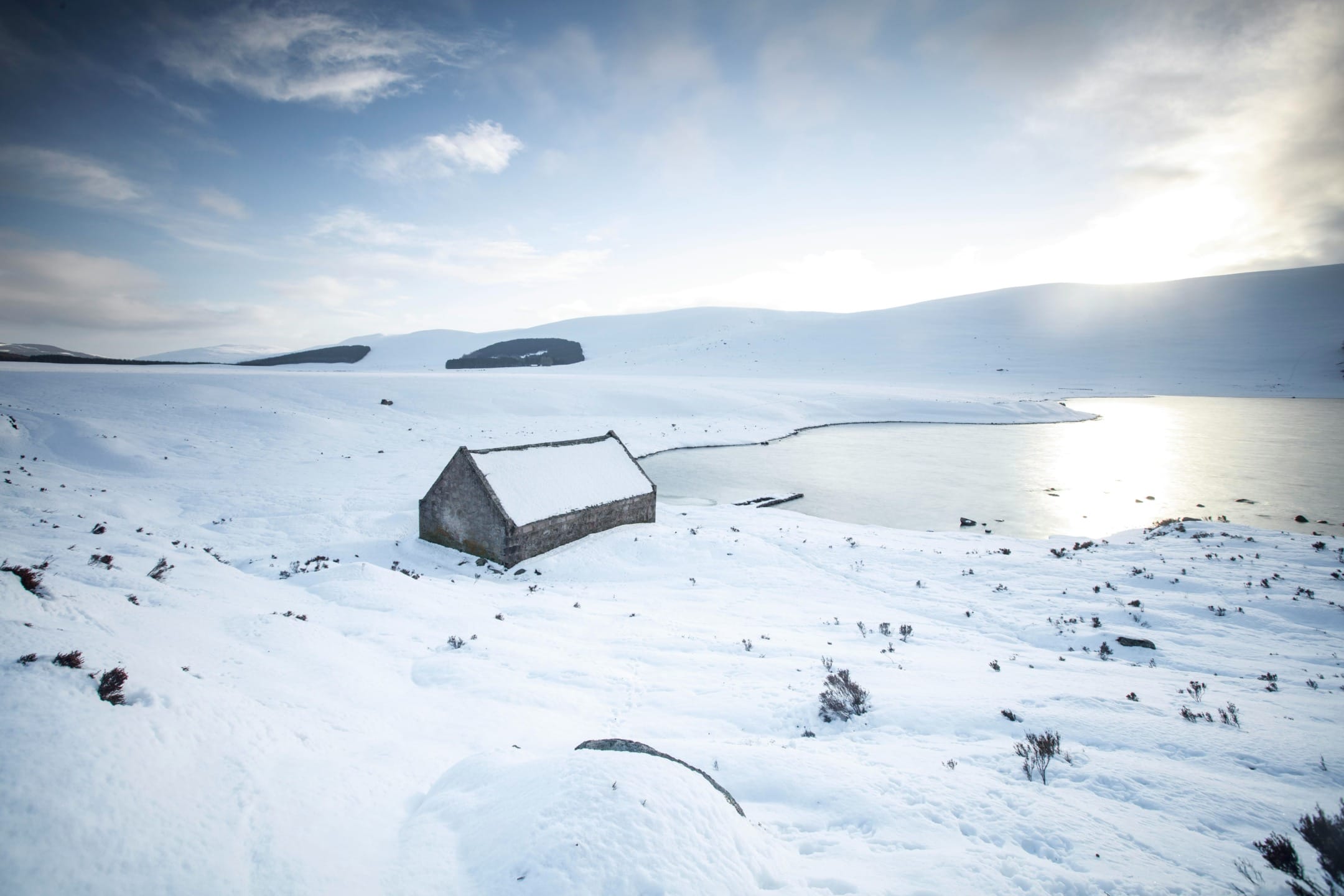 Snowy landscape with a small stone cabin near a frozen lake and mountains under a cloudy sky.